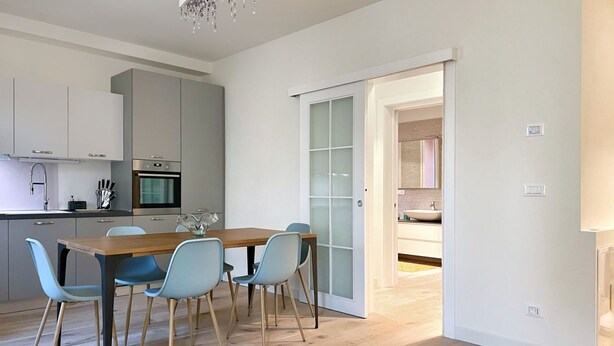 A modern kitchen with a dining area, featuring a wooden table and light blue chairs, a chandelier, and a sliding glass door leading to another room. - UR-NEST San Giorgio - gorgeous flat (Bologna)
