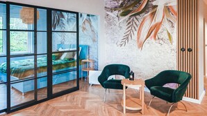 A modern room with a large window, a bed, two green chairs, a small table, and a wall with a leaf pattern. - Awesome apartment in Stezyca with sauna (Stezyca)