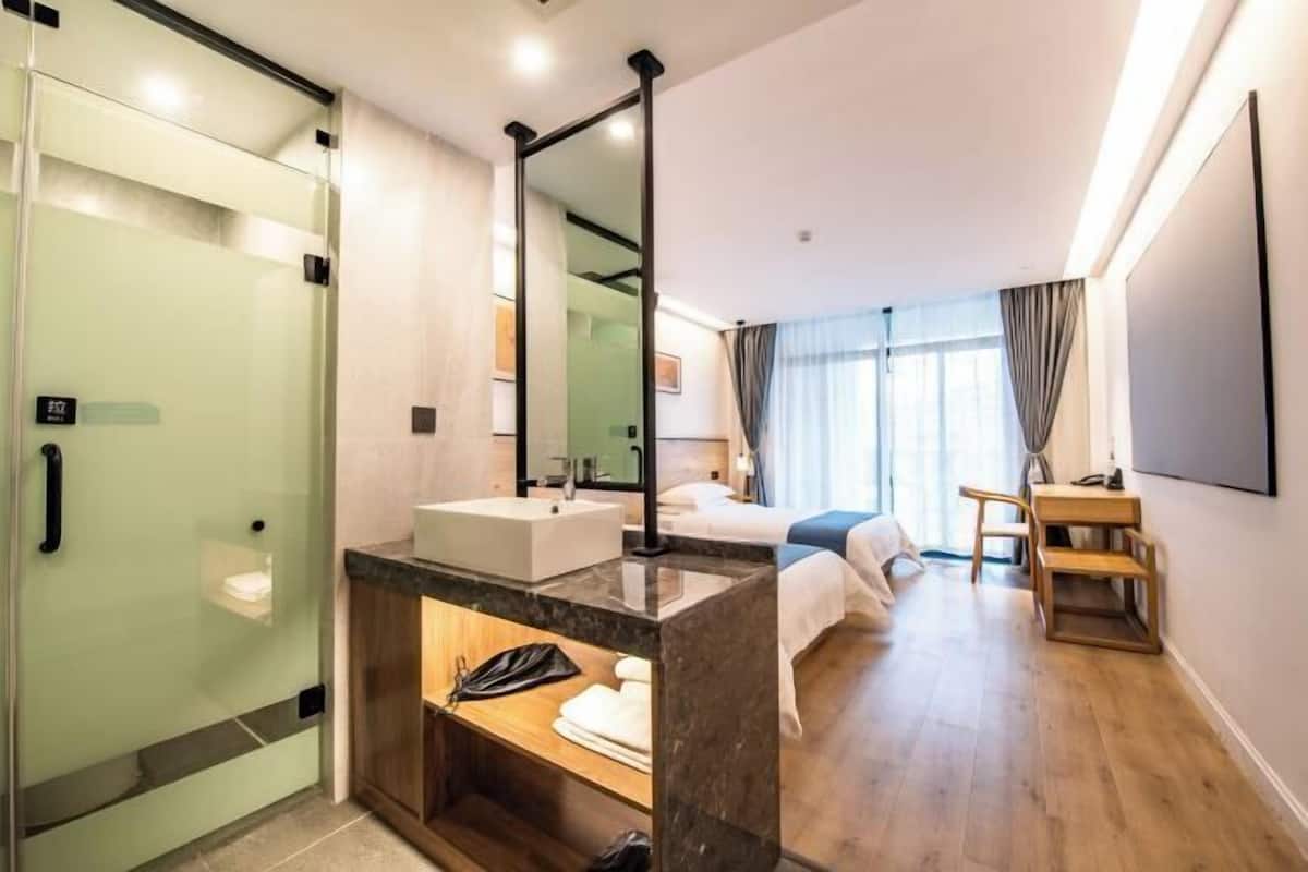 Habitación de hotel con ducha envuelta en vidrio, lavamanos, cama, escritorio y espejo grande.