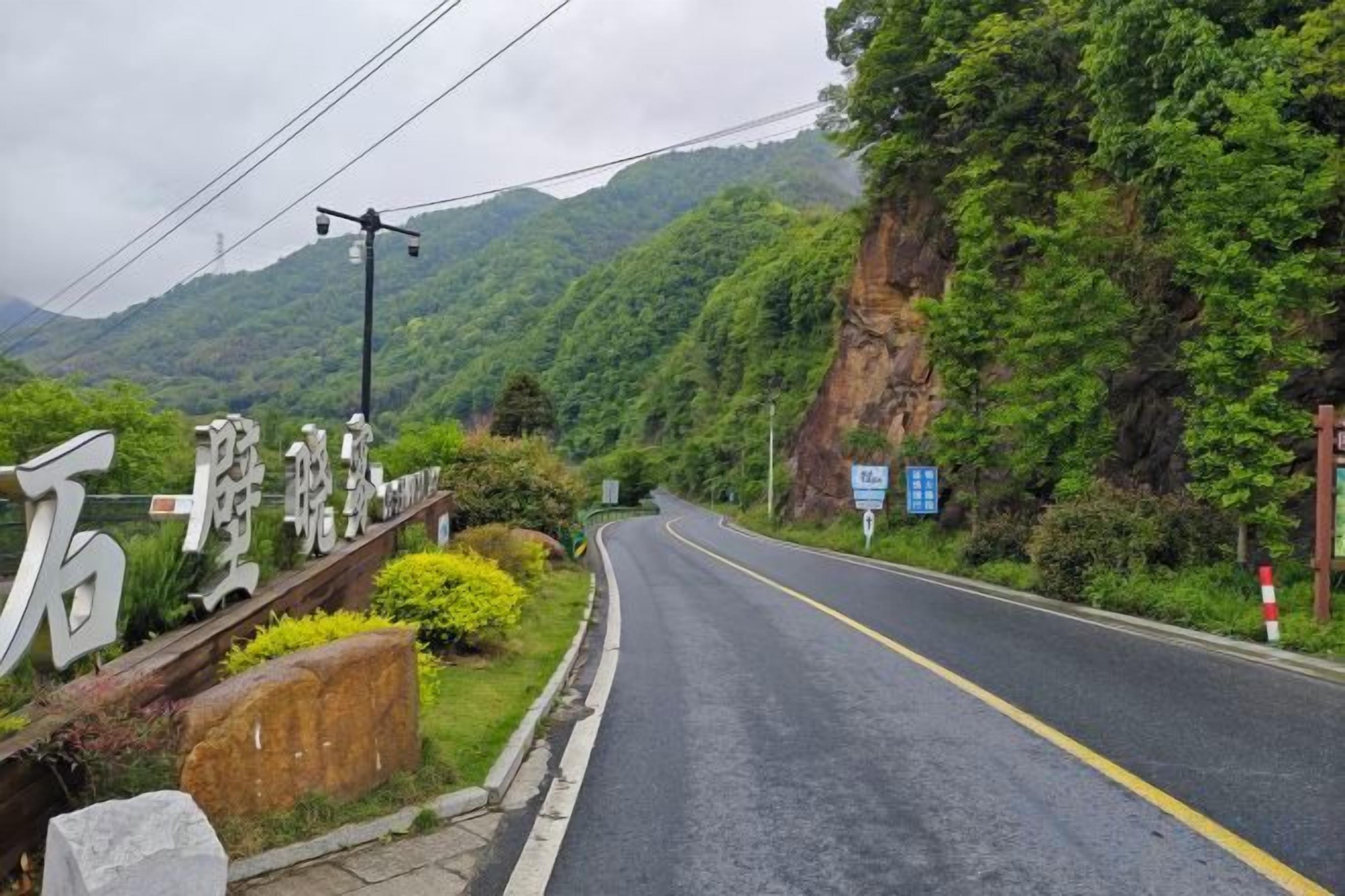 A road with a guardrail and a signpost on the left, surrounded by greenery and mountains.