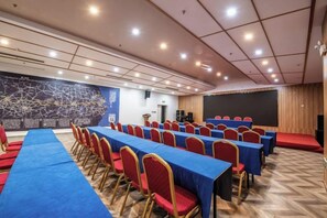 Un salón de conferencias con filas de mesas y sillas, un escenario y una pantalla de proyección.