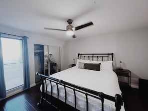 Um quarto com cama, ventoinha no teto, mesa de cabeceira, guarda-roupa e janela com cortinas.