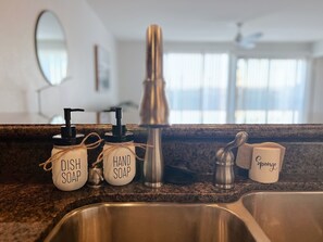 Uma bancada de cozinha com dosadores de sabão identificados como 'líquido para lavar louça' e 'sabão para as mãos', uma torneira de aço inoxidável e uma caneca com a inscrição 'esponja'.