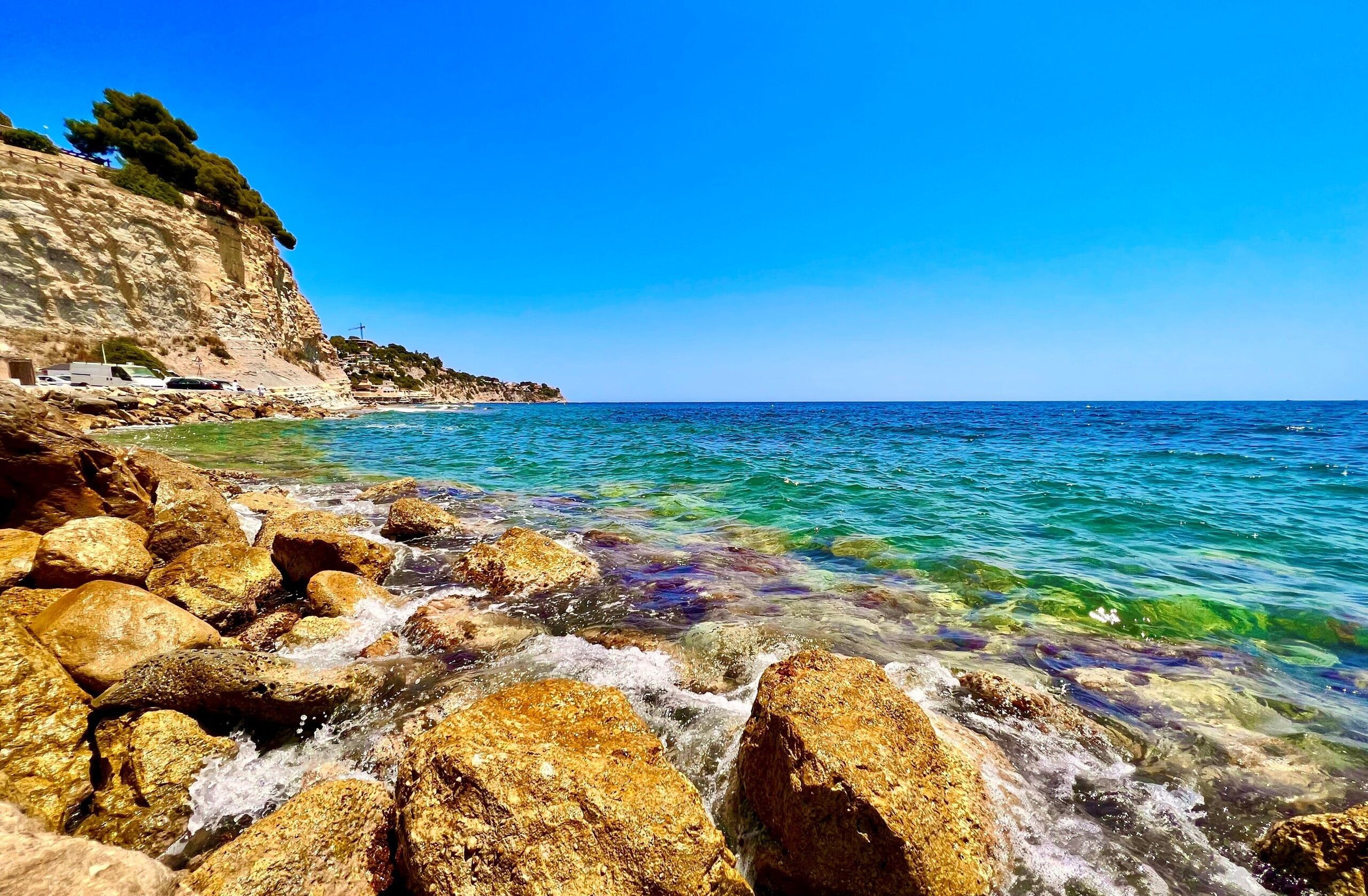 A rocky shoreline with clear blue water and a cliff with vegetation.