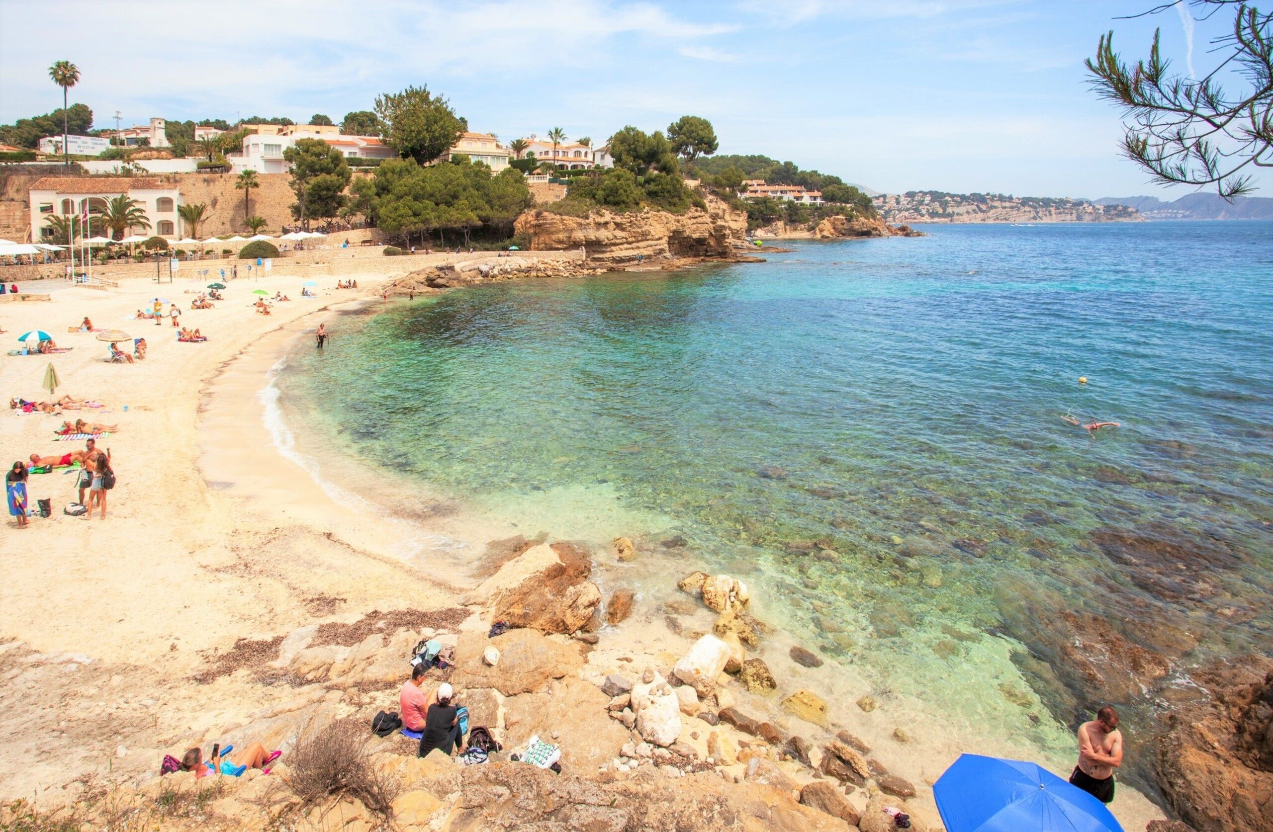 A coastal beach with clear waters, rocky areas, and buildings in the background.