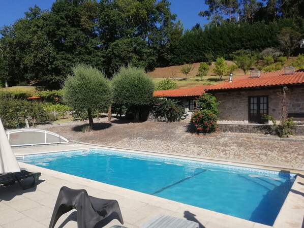 Une piscine bordée de pierres, un bâtiment en pierre avec un toit rouge et une végétation luxuriante tout autour.
