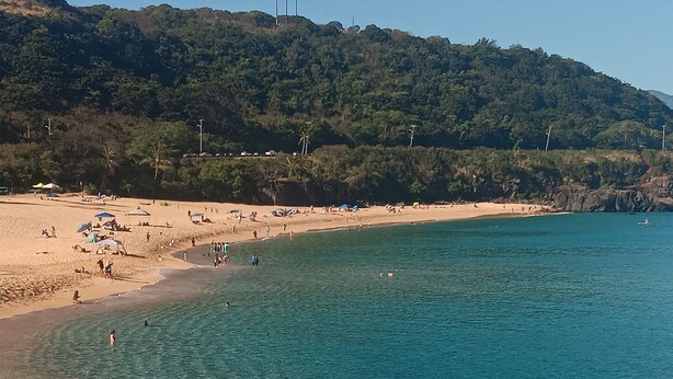 Une plage à l’eau turquoise et limpide, une rive sablonneuse et, en arrière-plan, une colline boisée.