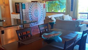Un coin repas avec une table en bois, des chaises et un bol décoratif. On aperçoit un canapé et une fenêtre avec des rideaux à l’arrière-plan.