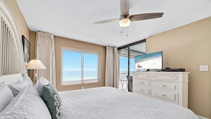 Un dormitorio con una cama grande, un ventilador de techo, un televisor sobre una cómoda y una ventana con vistas al cielo.