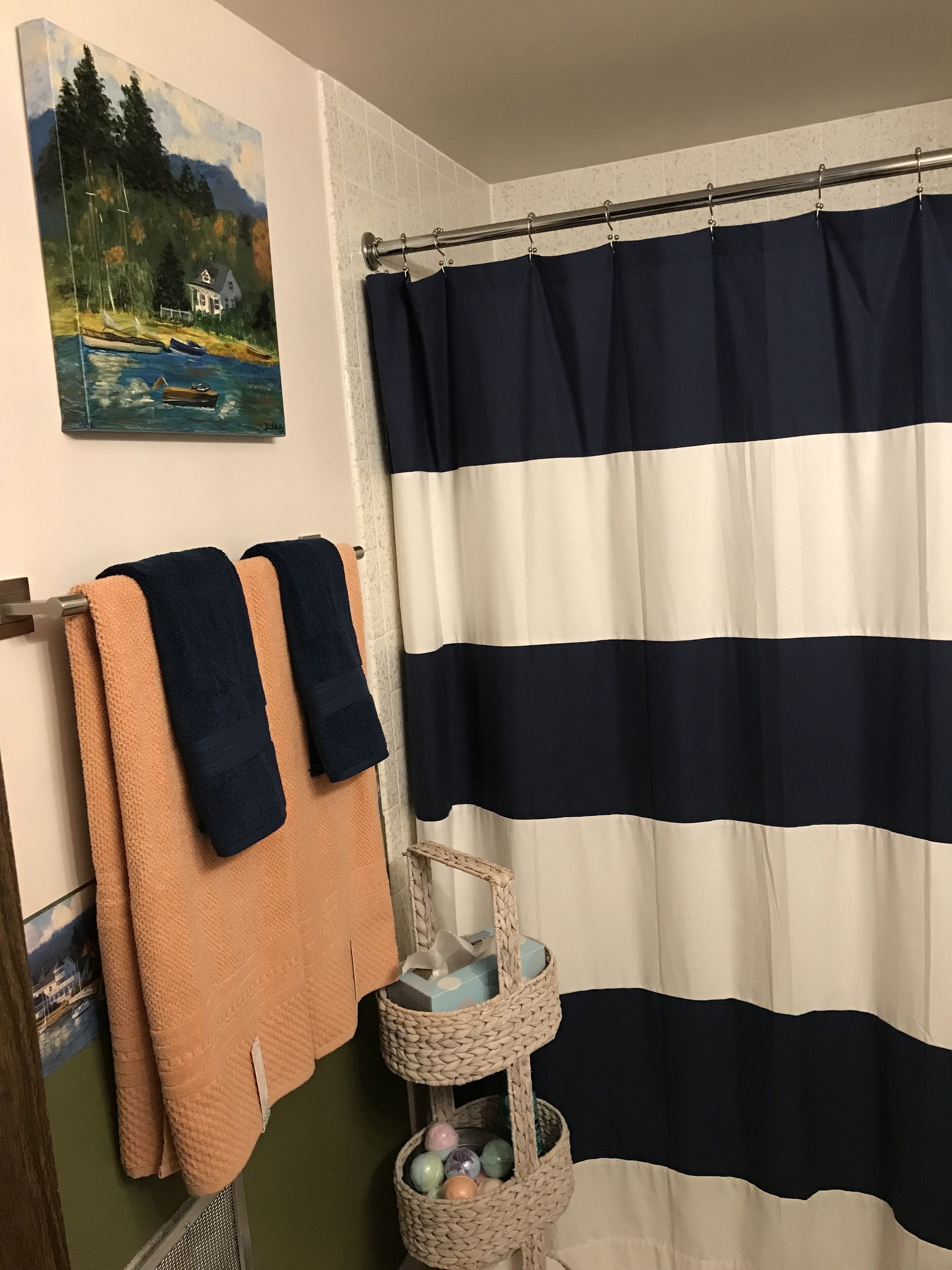 A bathroom with a striped shower curtain, a painting of a house by a lake, and a woven basket with decorative items.