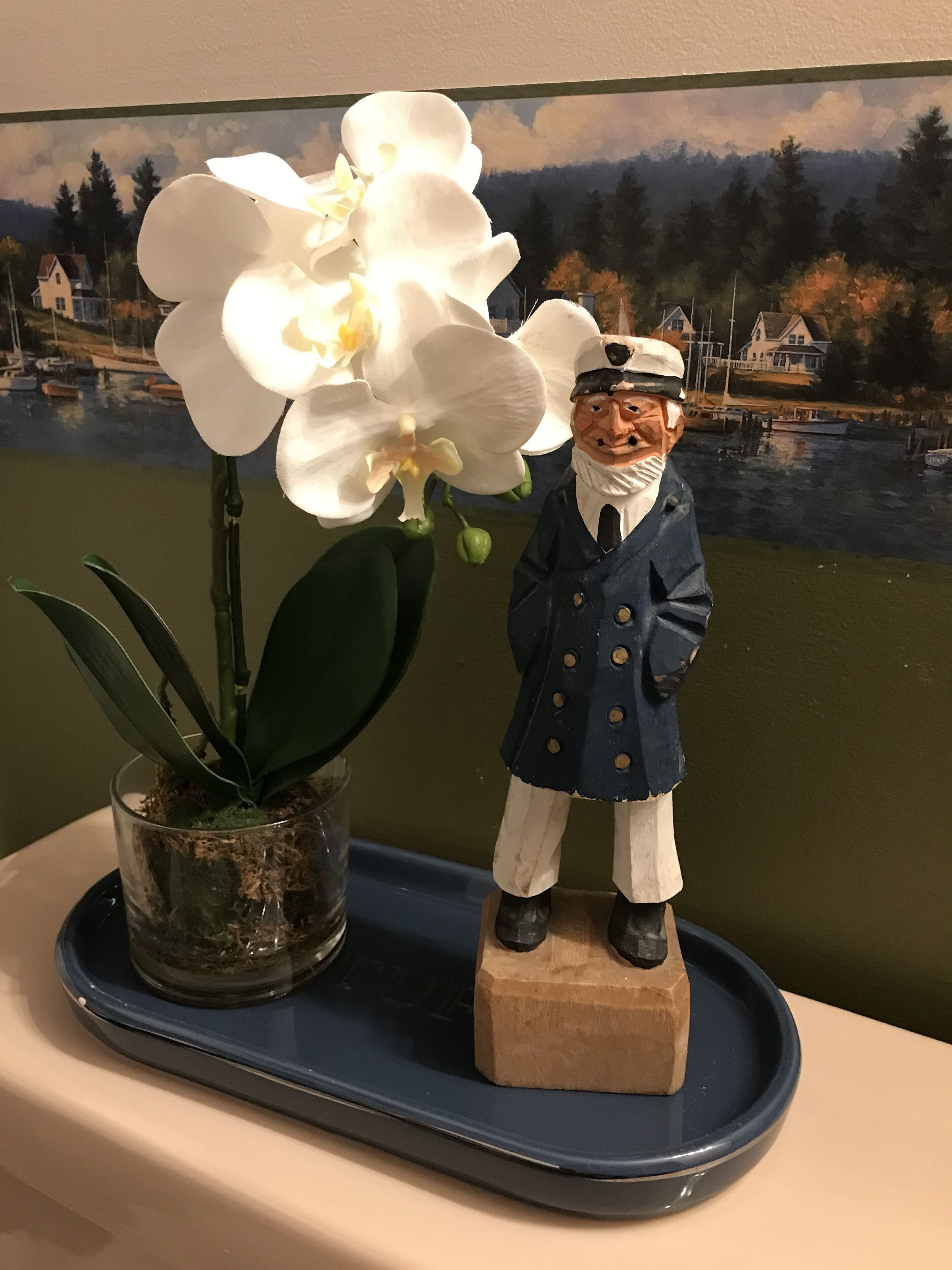 Une statuette représentant un marin manœuvrant une roue de navire, une orchidée en pot et un tableau représentant un paysage côtier.