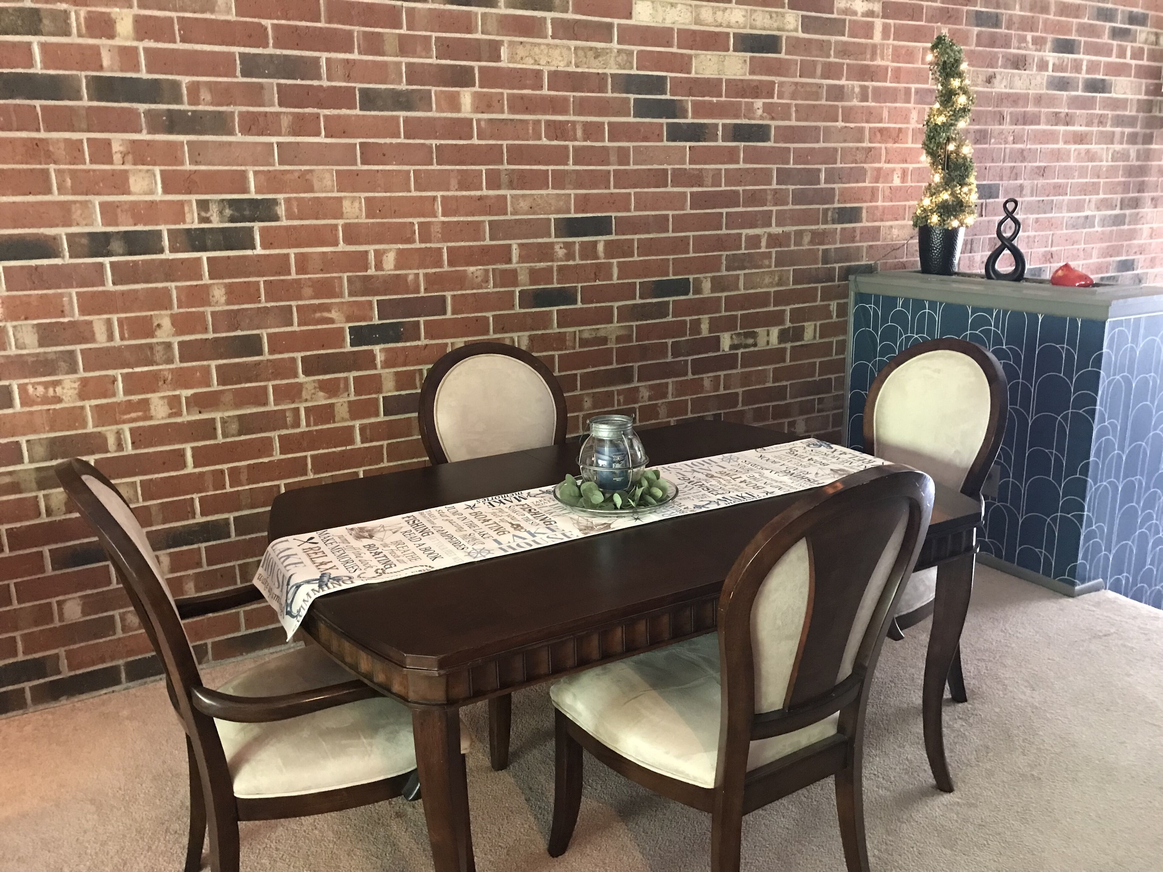 A dining table with four chairs, a decorative table runner, a small potted plant, and a candle holder.