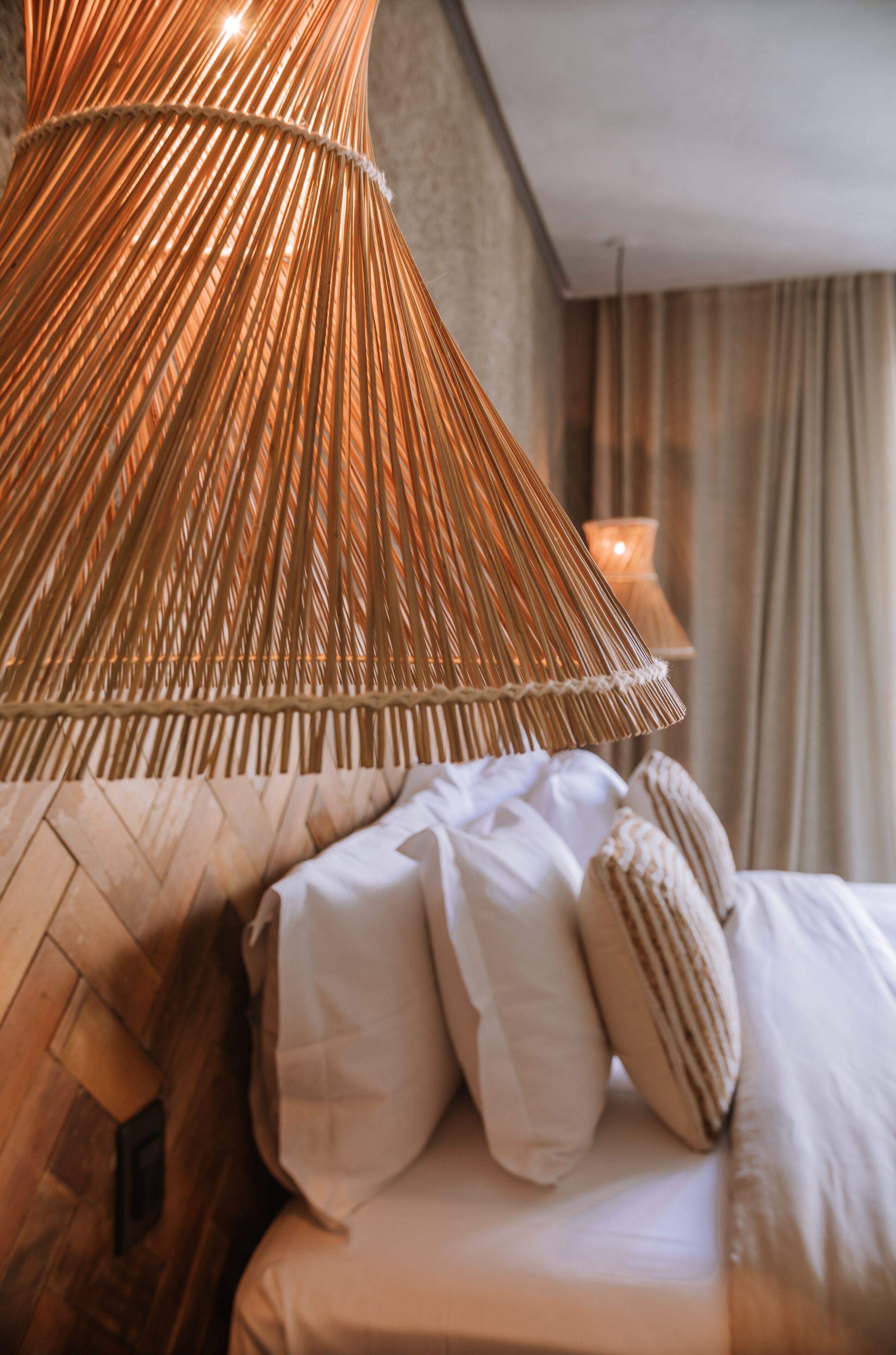Una cama con ropa blanca y almohadas a rayas, una lámpara de ratán trenzado y una vela al fondo.