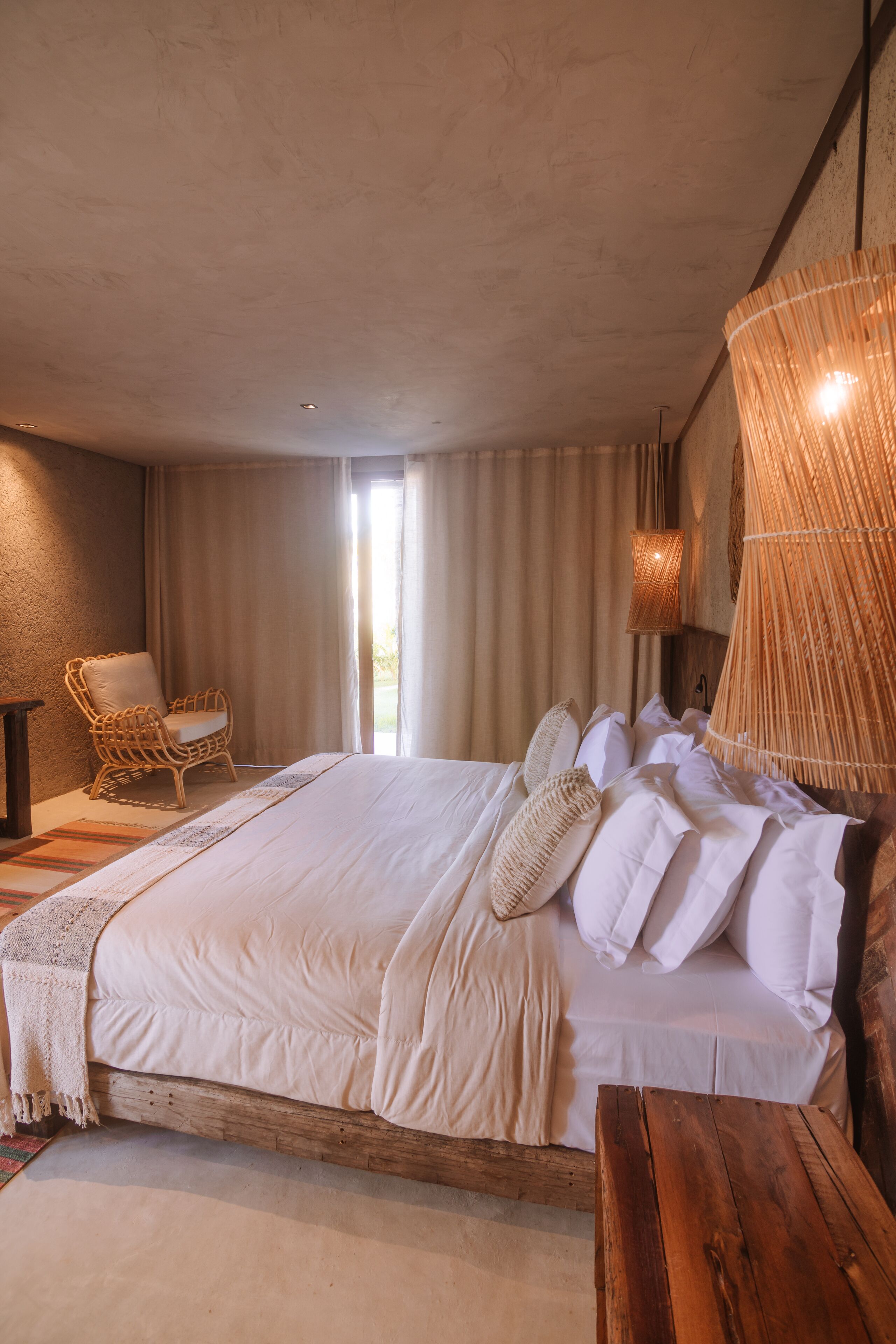 Quarto com uma cama grande, uma cadeira de vime, um banco de madeira e um lustre tecido grande.