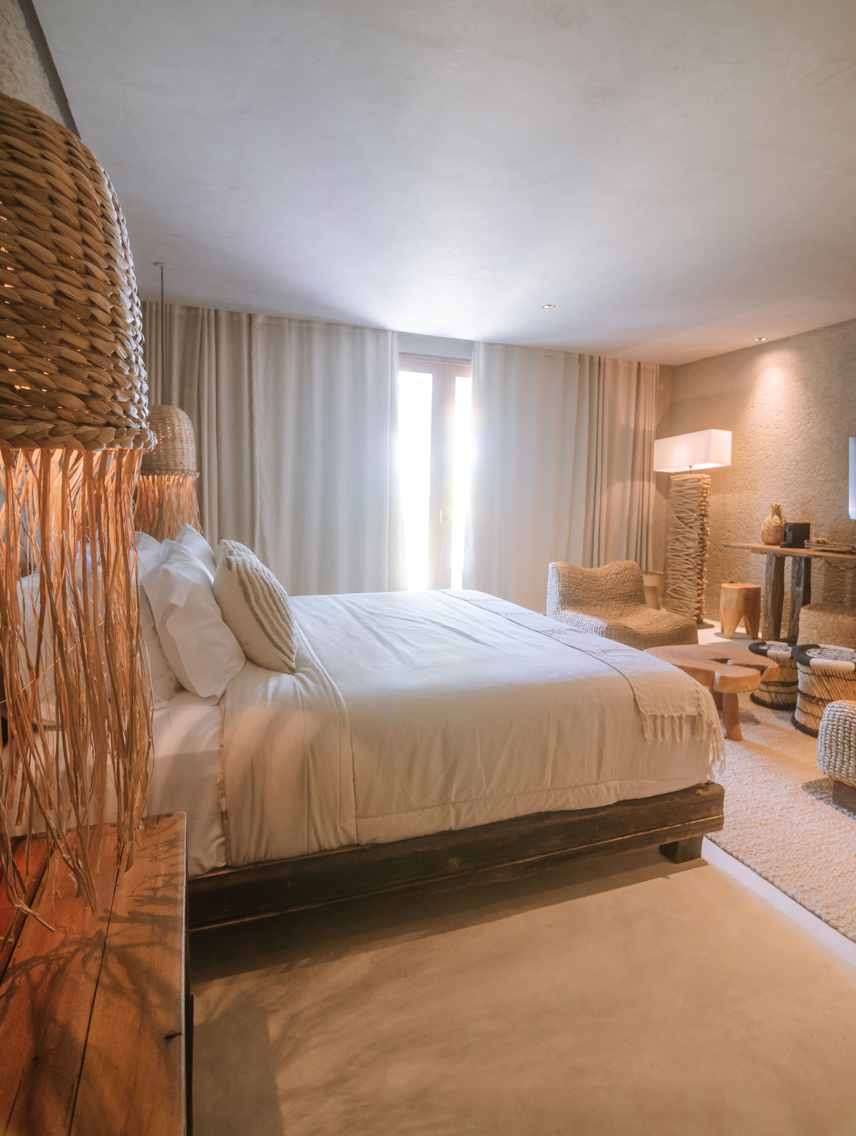A bedroom with a large bed, a woven headboard, a bedside table, a lamp, and a view of the outdoors through a window.