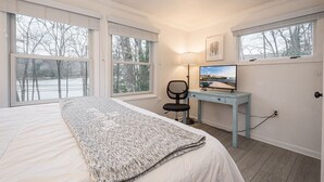 Een slaapkamer met een groot bed, een bureau met stoel, een televisie en twee ramen met jaloezieën.
