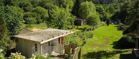 A rustic cabin with a gravel roof, surrounded by lush greenery and a hilly backdrop.
