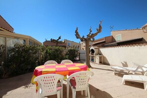 A patio area with a table covered by a colorful tablecloth, surrounded by white plastic chairs.