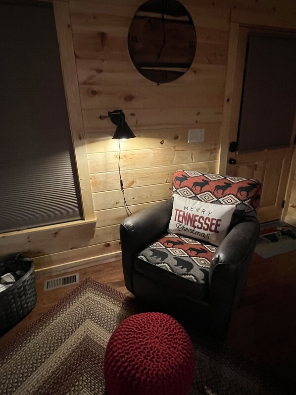 Une pièce chaleureuse avec un fauteuil en cuir, un pouf rouge, une horloge murale et une fenêtre avec des stores.