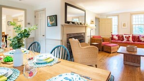 Un comedor con una mesa de madera lista para una comida, una chimenea y una zona de estar con un sofá y sillones.