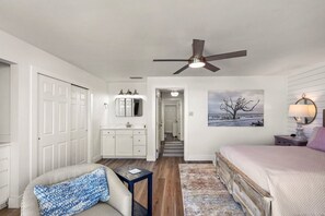 Une chambre à coucher comprenant un lit, un ventilateur de plafond, une table de chevet, une lampe, une commode, un miroir, une porte et un couloir menant à une autre pièce.