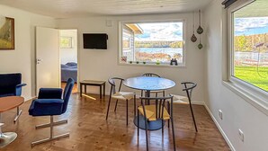 Ein Zimmer mit Holzboden, einem runden Tisch mit Stühlen, einem an der Wand befestigten Fernseher und einem Fenster mit Blick auf Bäume und ein Gebäude.