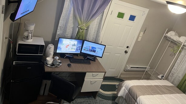 A bedroom with a desk, two monitors, a coffee maker, and a bed.