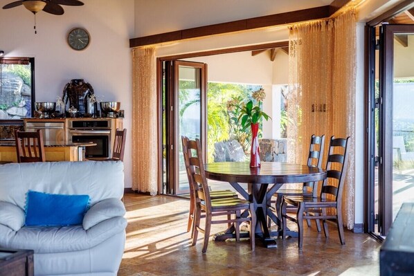 Un comedor con una mesa redonda y sillas, una zona de cocina con electrodomésticos, y vistas a zonas verdes a través de amplios ventanales.