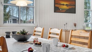 Ruokailutila, jossa on neljälle katettu pöytä. Pöydällä on kukkakuviollinen pöytäliina, kaksi valkoista tuolia, puinen penkki, kulho salaattia, lautanen mansikoita ja muffinsseja sekä valkoinen kannu.