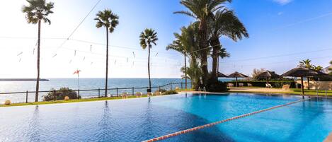 A clear blue swimming pool with a view of the sea, surrounded by palm trees and lounge chairs.