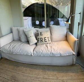 Un sofá color beige con varios cojines, incluyendo uno con el texto "BE FREE", situado cerca de una ventana que ofrece vistas a árboles y a una persona tomando una fotografía.