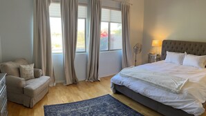 Une chambre à coucher avec un lit, une table de chevet, une lampe, un ventilateur, une fenêtre avec des rideaux, un canapé avec des coussins et un tapis bleu.