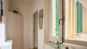 Un pasillo estrecho con una puerta que da a una habitación, una ventana con contraventanas verdes y un radiador con plantas en macetas.