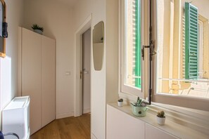 Ein schmaler Korridor mit einer zur einem Raum führenden Tür, einem Fenster mit grünen Fensterläden und einem Heizkörper mit Blumentöpfen.