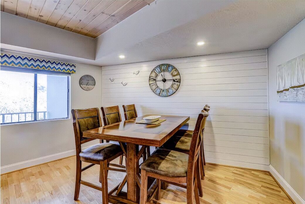 A dining area with a wooden table and chairs, a large wall clock, and a window with a view.