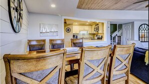A dining area with a wooden table and chairs, a clock on the wall, and a view into a kitchen area.