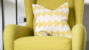 Ein senffarbener Sessel mit einem Kissen in geometrischem Muster, eine Stehlampe und eine Zeitschrift auf einem Tisch.