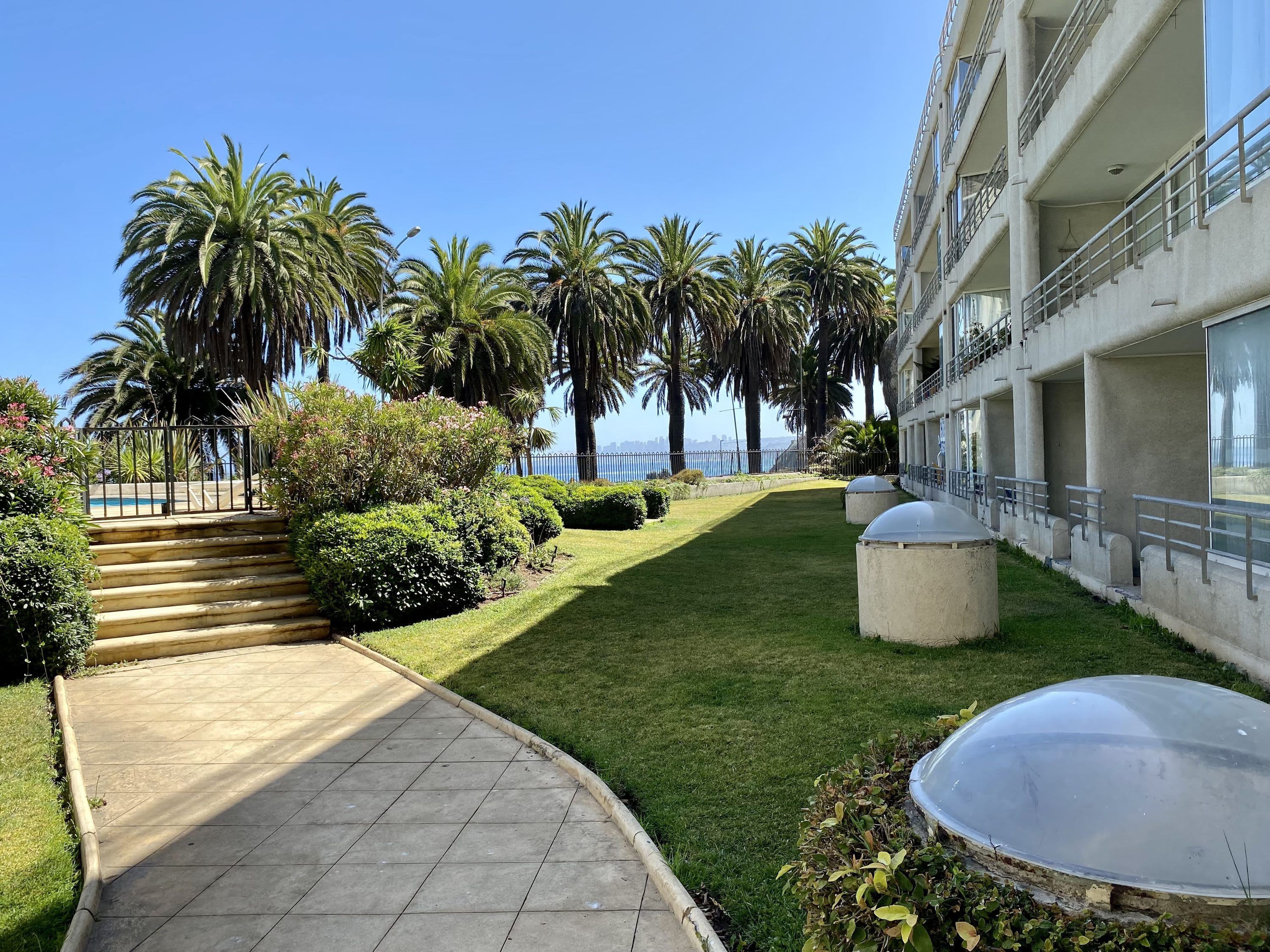 A pathway leading to a building with a view of the sea, surrounded by palm trees and well-maintained grass.