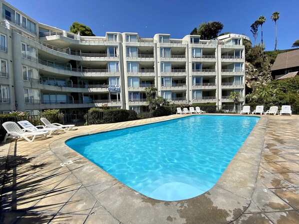 A swimming pool with lounge chairs, a multi-story building with balconies, and a clear blue sky.