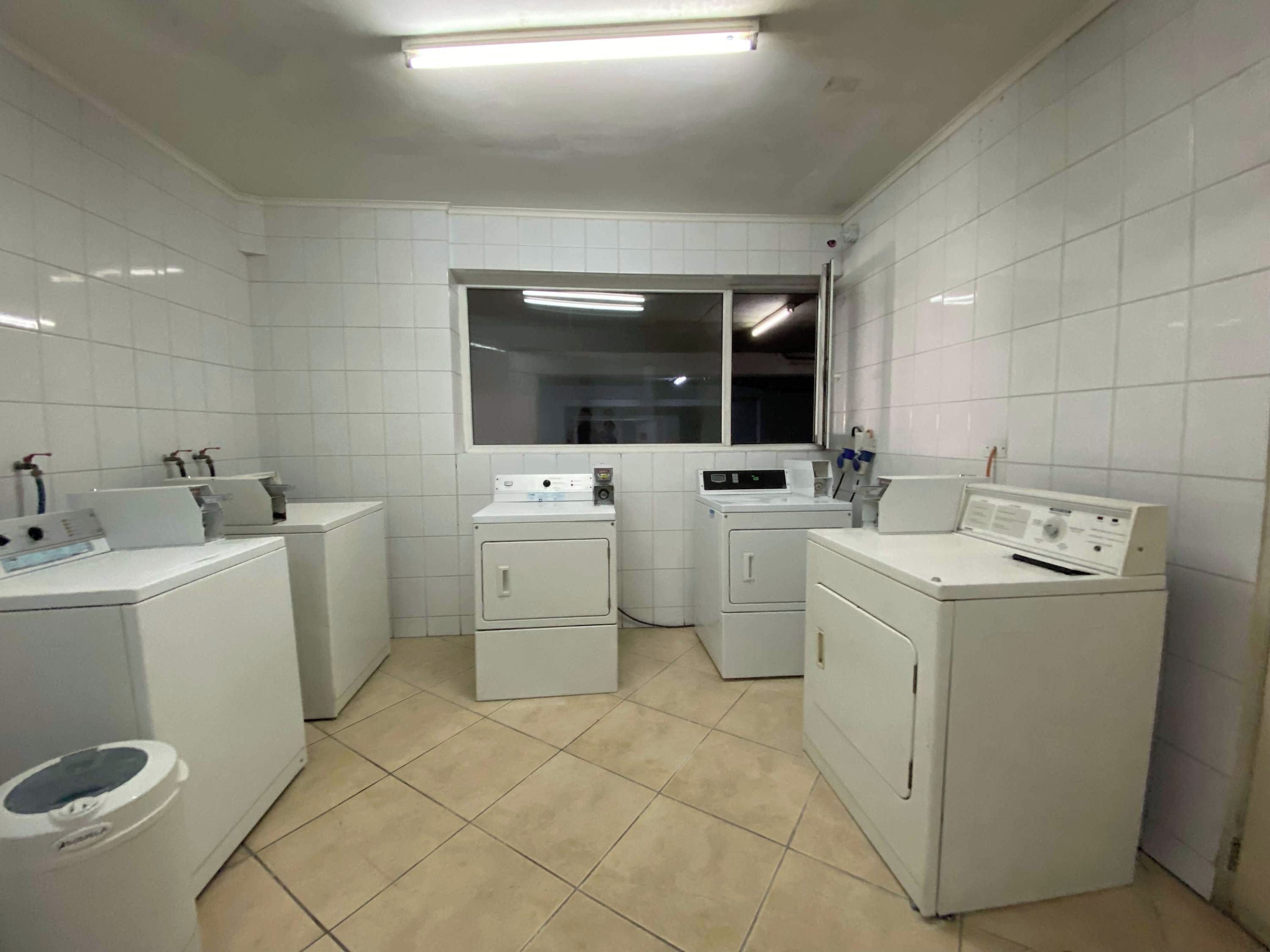 A laundry room with white tiled walls, a window, and multiple washing machines.