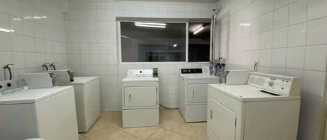 A laundry room with white tiled walls, a window, and multiple washing machines.