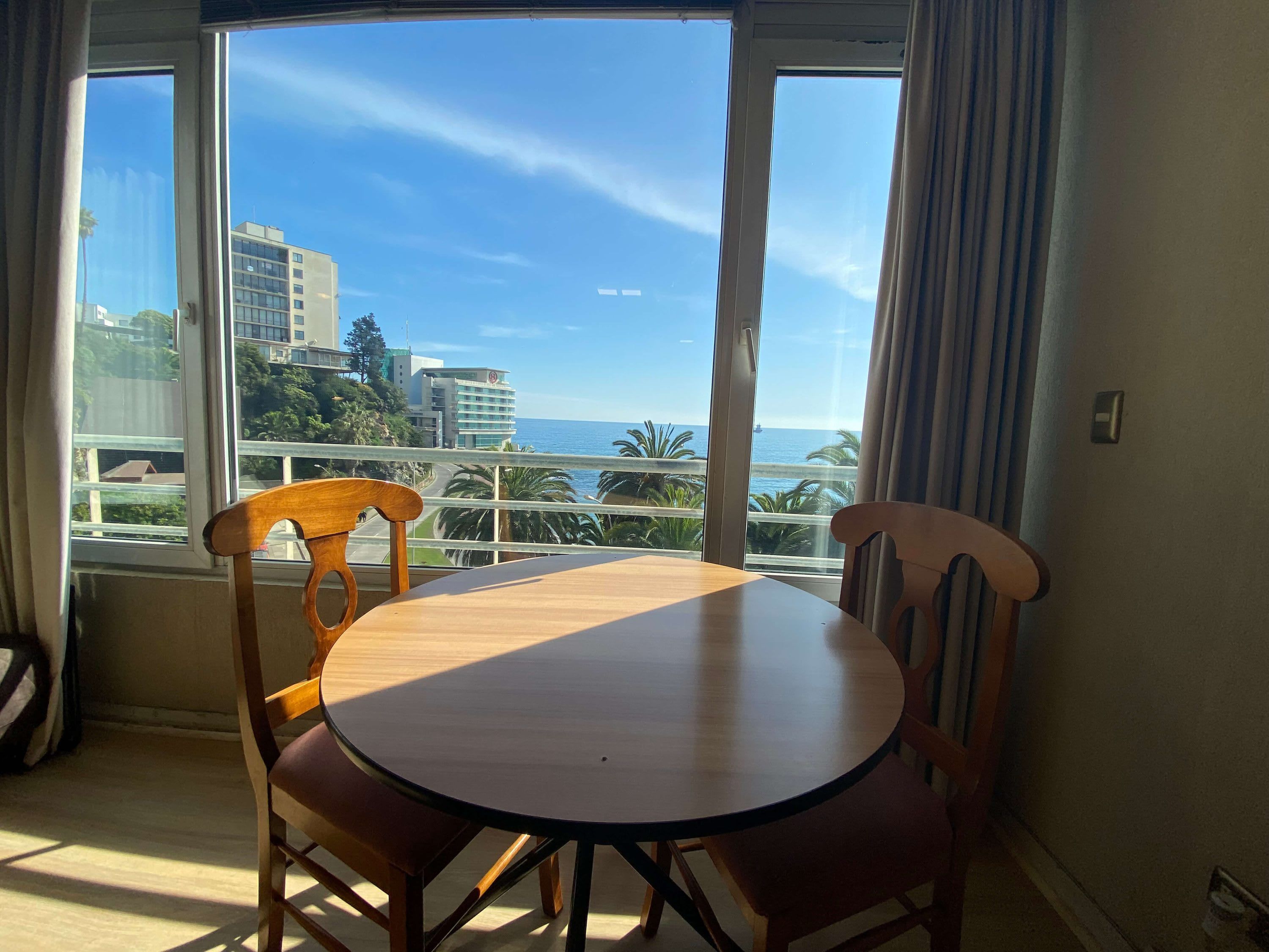 A room with a round table and two chairs, a window with a view of the sea and buildings, and a curtain on the window.