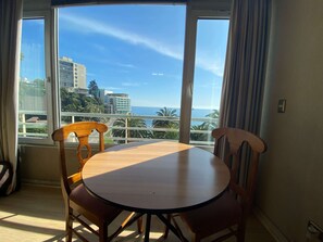 Una habitación con una mesa redonda y dos sillas, un ventanal con vistas al mar y edificios, y una cortina en la ventana.