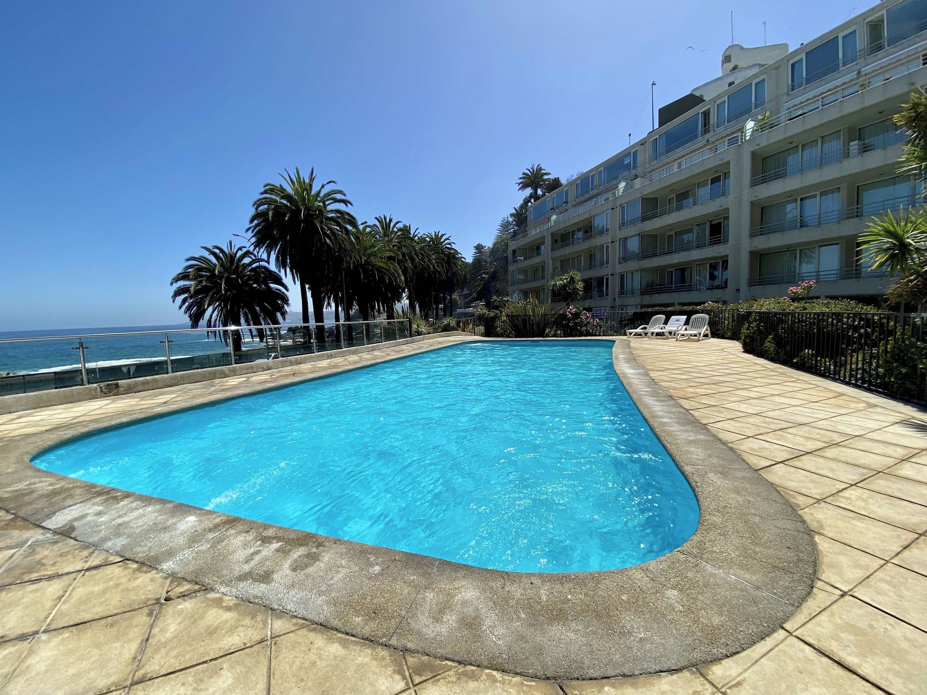 A swimming pool with a curved edge, surrounded by a tiled deck, palm trees, and a building with balconies.