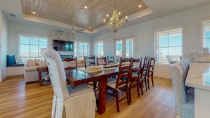 A spacious dining area with a wooden table and chairs, a chandelier, and large windows offering views of the outdoors. - Gulf Views, Private Heated Pool, Golf Cart Included, Boardwalk to the Beach (Port Aransas)