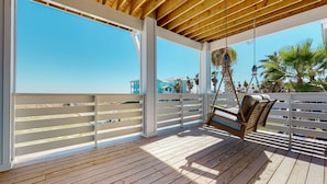 A wooden deck with a swing chair, white railings, and a view of buildings and palm trees. - Gulf Views, Private Heated Pool, Golf Cart Included, Boardwalk to the Beach (Port Aransas)