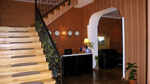 A wooden staircase with a metal railing, a reception desk with a lamp, clocks, and flags. - Darchi Hotel (Tbilisi)