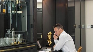 Un hombre sentado en un escritorio con una computadora portátil, un mueble de vidrio con botellas y un espejo grande.
