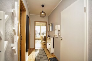 A hallway with a wooden door, a white wall, a ceiling light, and a view into a room with a desk and chair.