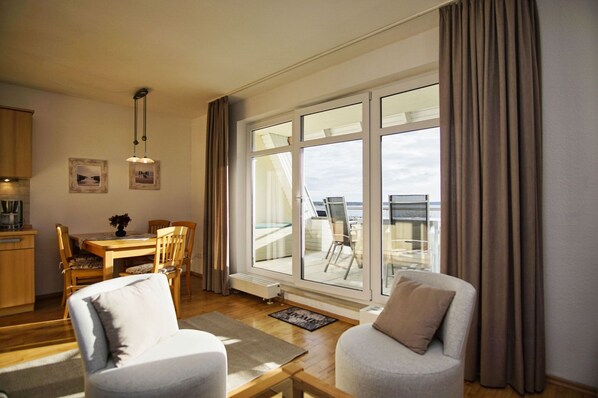 A modern living room with a dining area, large windows with curtains, and a view of the sea.